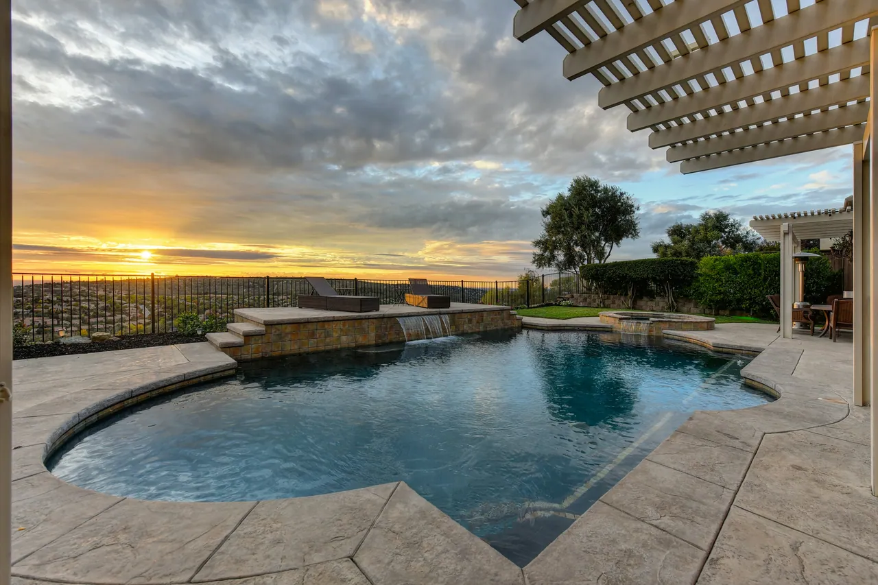A backyard pool with a stone water feature under a pergola at sunset.
