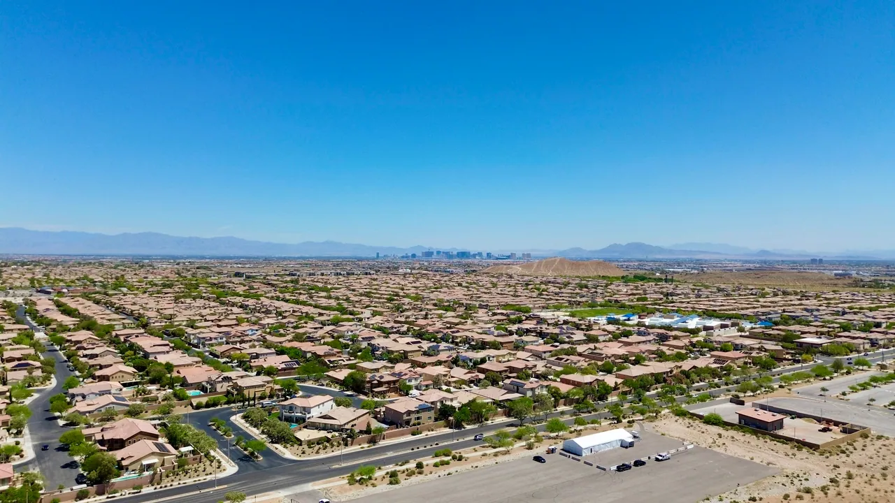 Drone view across a Las Vegas desert neighborhood of stucco homes under a clear sky, with the city and mountains on the horizon.